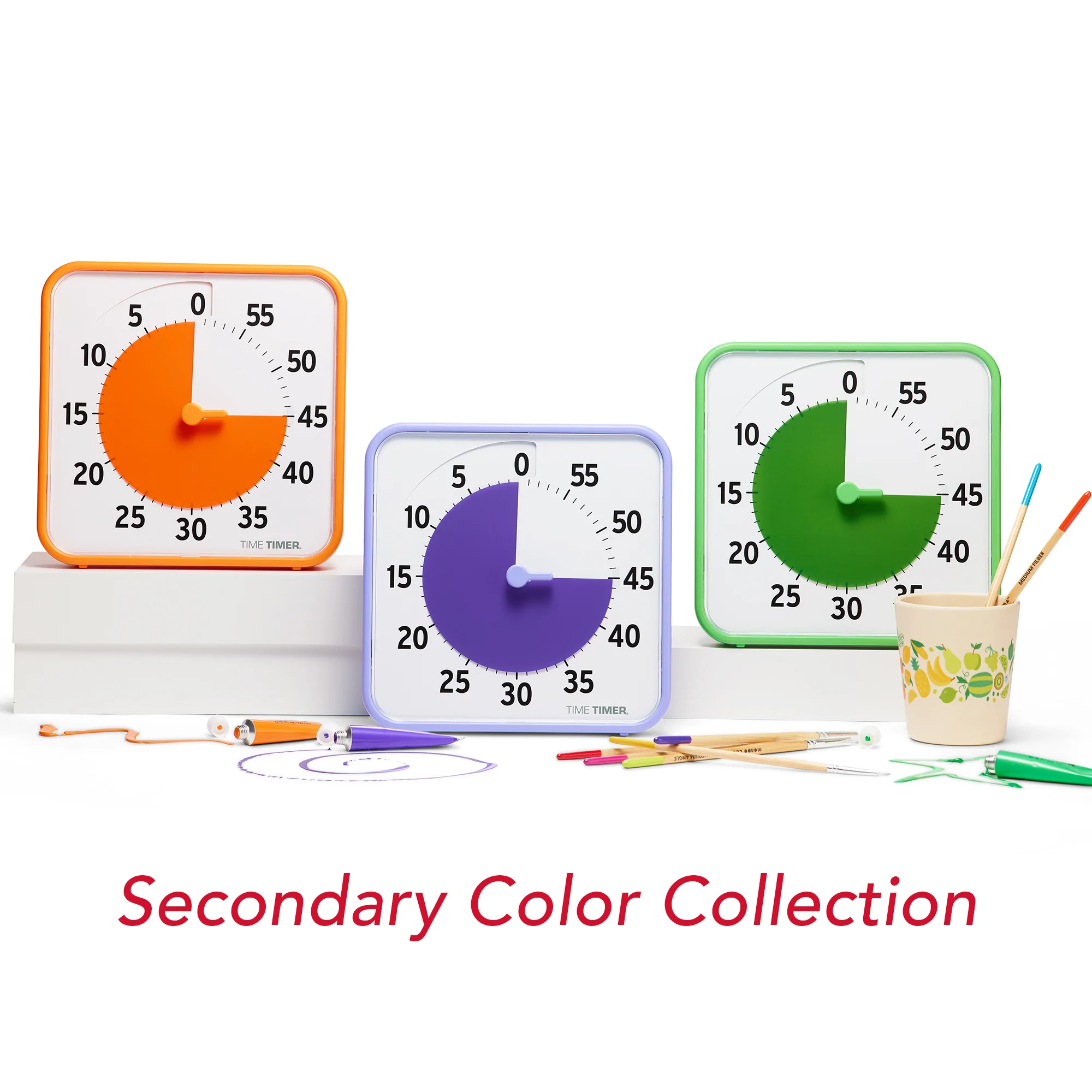 Oranž, lilla ja roheline Time Timer laual kõrvuti, lilla teistest madalama, taimeite ees pliiatsid ja värvituubid ning pliiatsitops kahe pliiatsiga. Pildi all ingliskeelne tekst "Secondary Color Collection"
