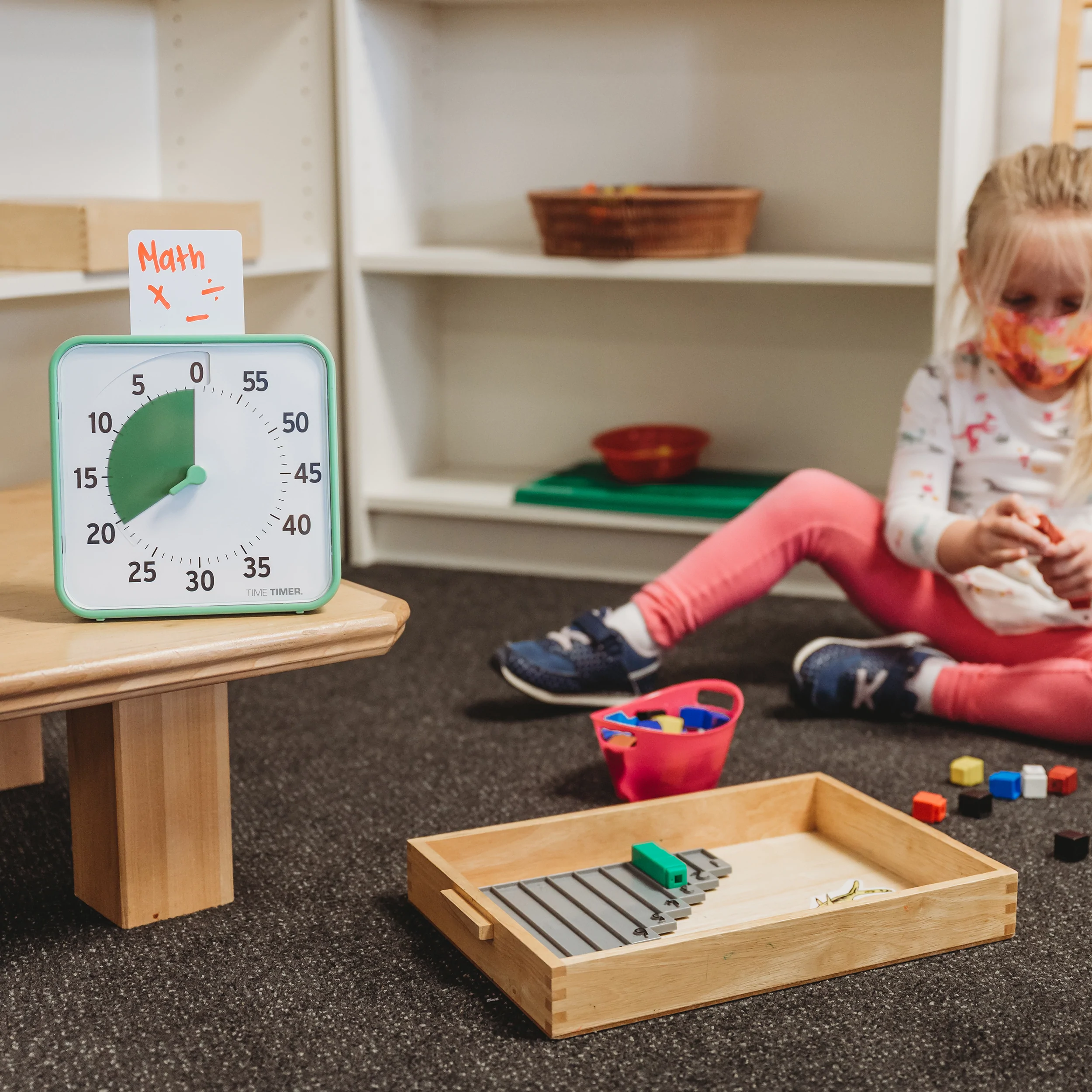 Roheline Time Timer väiksel laual, taimer ülaossa asetatud valge kaart, millele on inglise keeles kirjutatud "math" ehk matemaatika. Maas väike puidust kastike, mille sees ja ümber on mänguasjad, taamal väike blond, umbes 3-aastane tüdruk maskiga mängimas klotsidega.